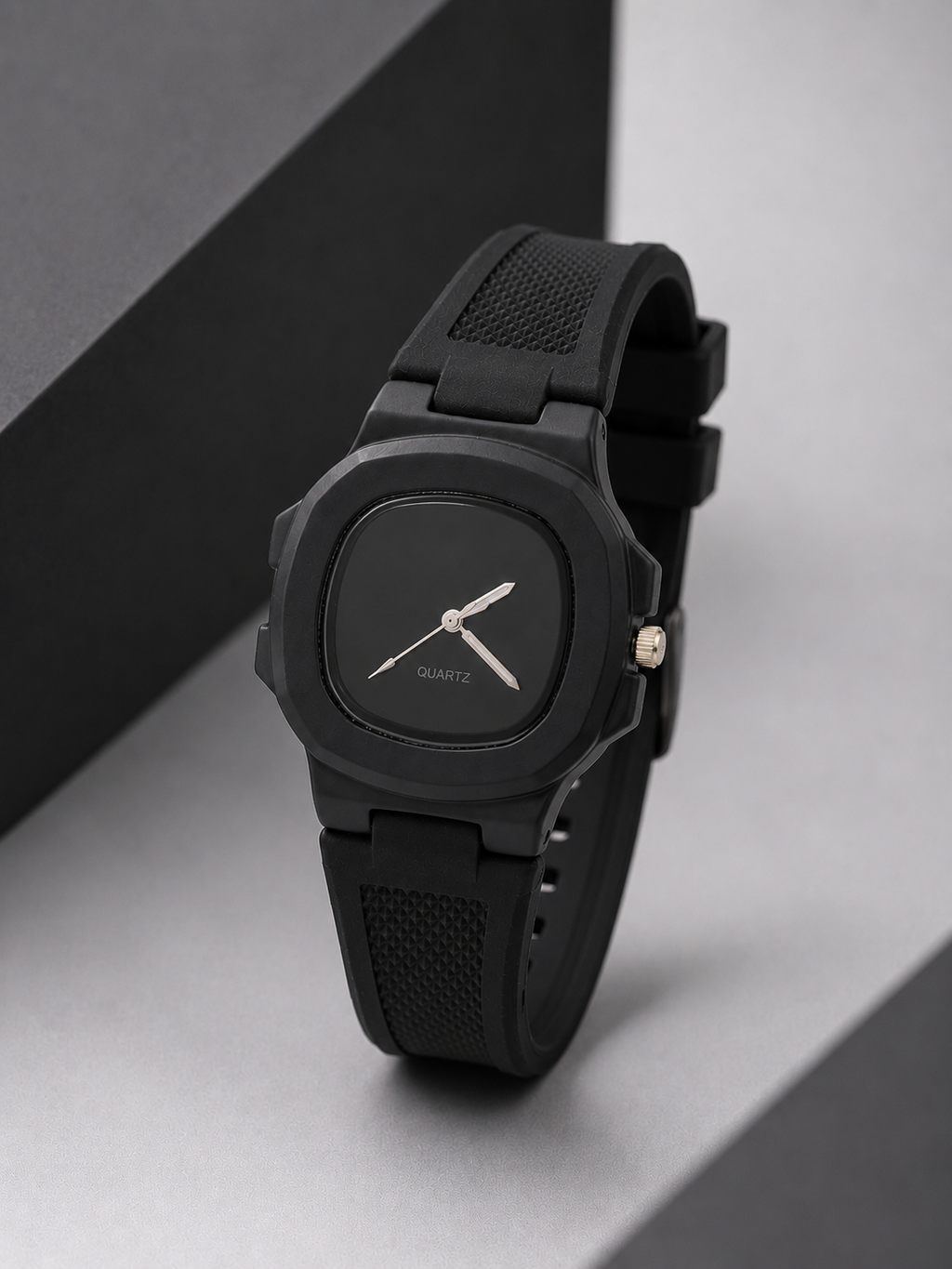 A minimalist black analog watch with a square case, black textured strap, and silver-tone hands, displayed on a gray surface with a black geometric prop.