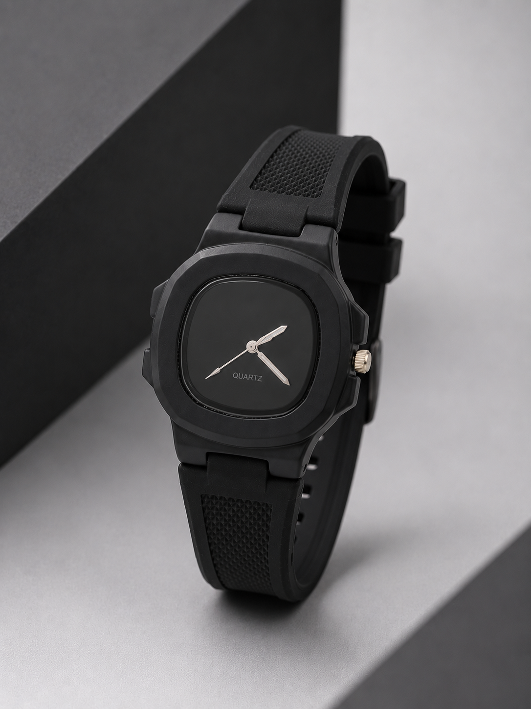 A minimalist black analog watch with a square case, black textured strap, and silver-tone hands, displayed on a gray surface with a black geometric prop.