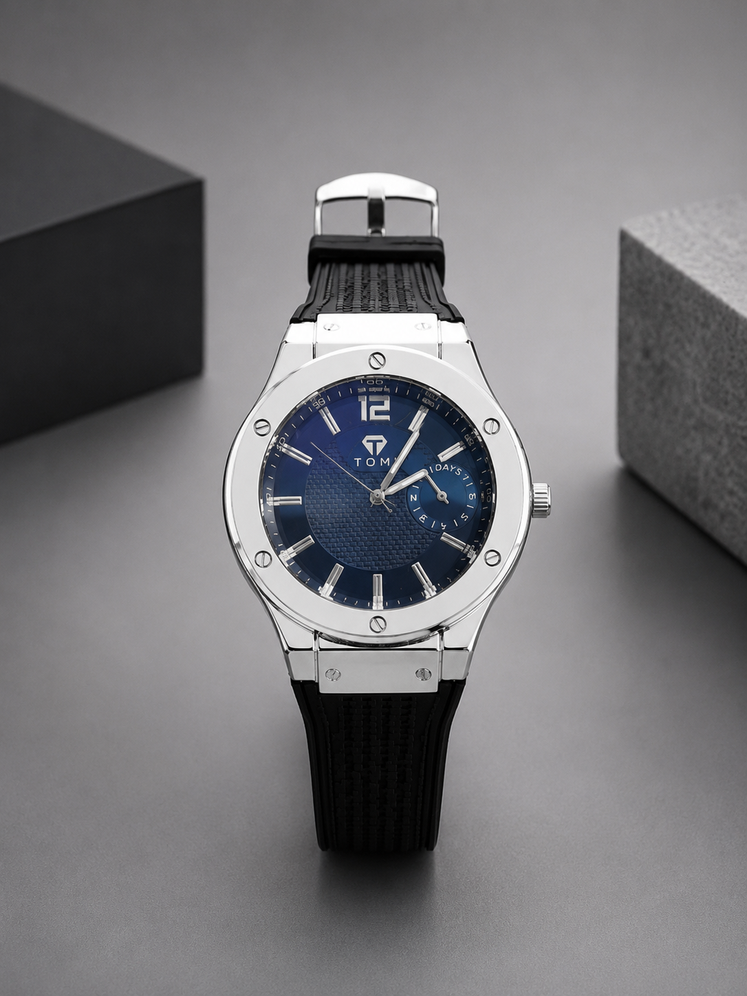A wristwatch with a silver-tone stainless steel case, a textured blue dial, and a black textured strap, displayed on a dark gray surface with two dark boxes in the background.