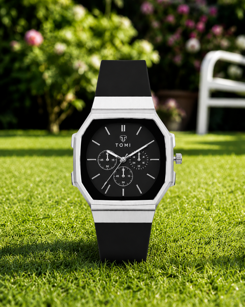 A TOMI watch with a black dial and black strap, set against a blurred green garden background.