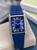 A rectangular analog watch with a blue strap, blue dial, and silver-tone case, resting on a beige surface with a blurred background.