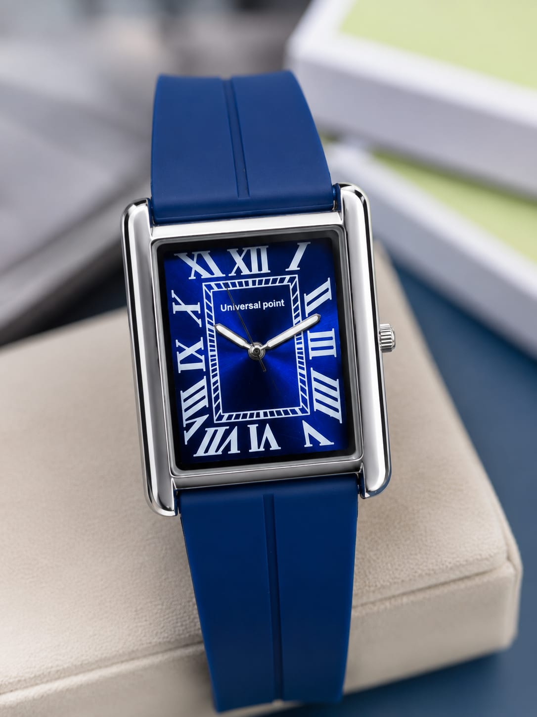 A rectangular analog watch with a blue strap, blue dial, and silver-tone case, resting on a beige surface with a blurred background.