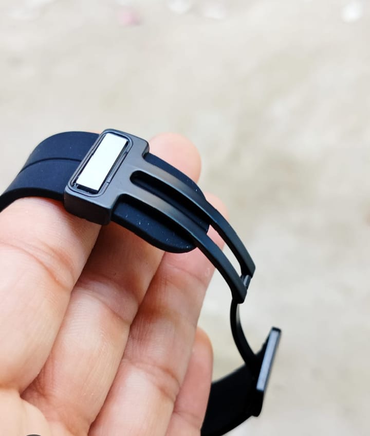 A close-up of a hand holding a black silicone watch band with a matte black metal spring-ring clasp and a small white rectangular accent.