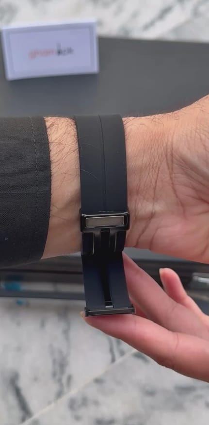A close-up of a black silicone watch band with a black metal buckle, shown being worn on a person's wrist and held by another hand.
